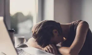 Zmęczona osoba z głową opartą na rękach przy laptopie, kubek kawy obok. Objawy neurastenii.