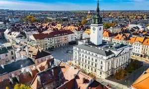 Widok z lotu ptaka na rynek z ratuszem i wieżą zegarową, otoczony kamienicami.