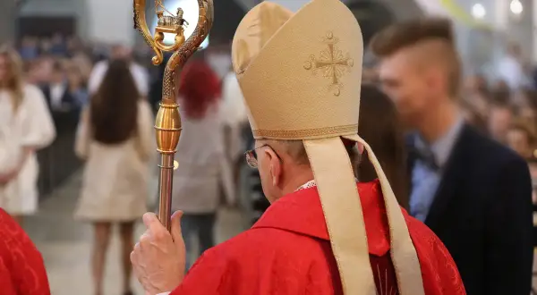 Biskup w czerwonej szacie i mitrze z krzyżem, trzyma pastorał ze złotym barankiem.