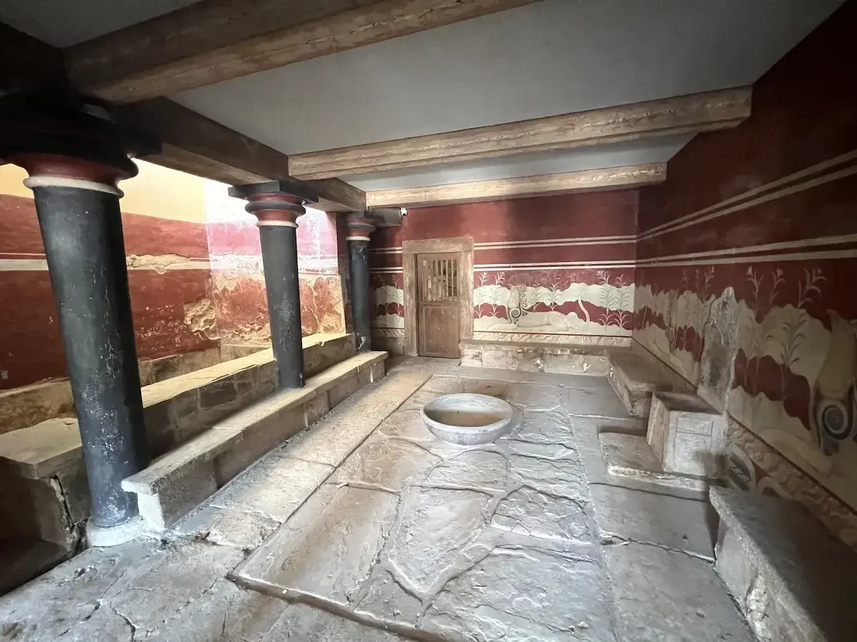 Wnętrze pałacu w Knossos z czarnymi kolumnami i freskami. Kamienna posadzka i kamienne ławy tworzą antyczną atmosferę.