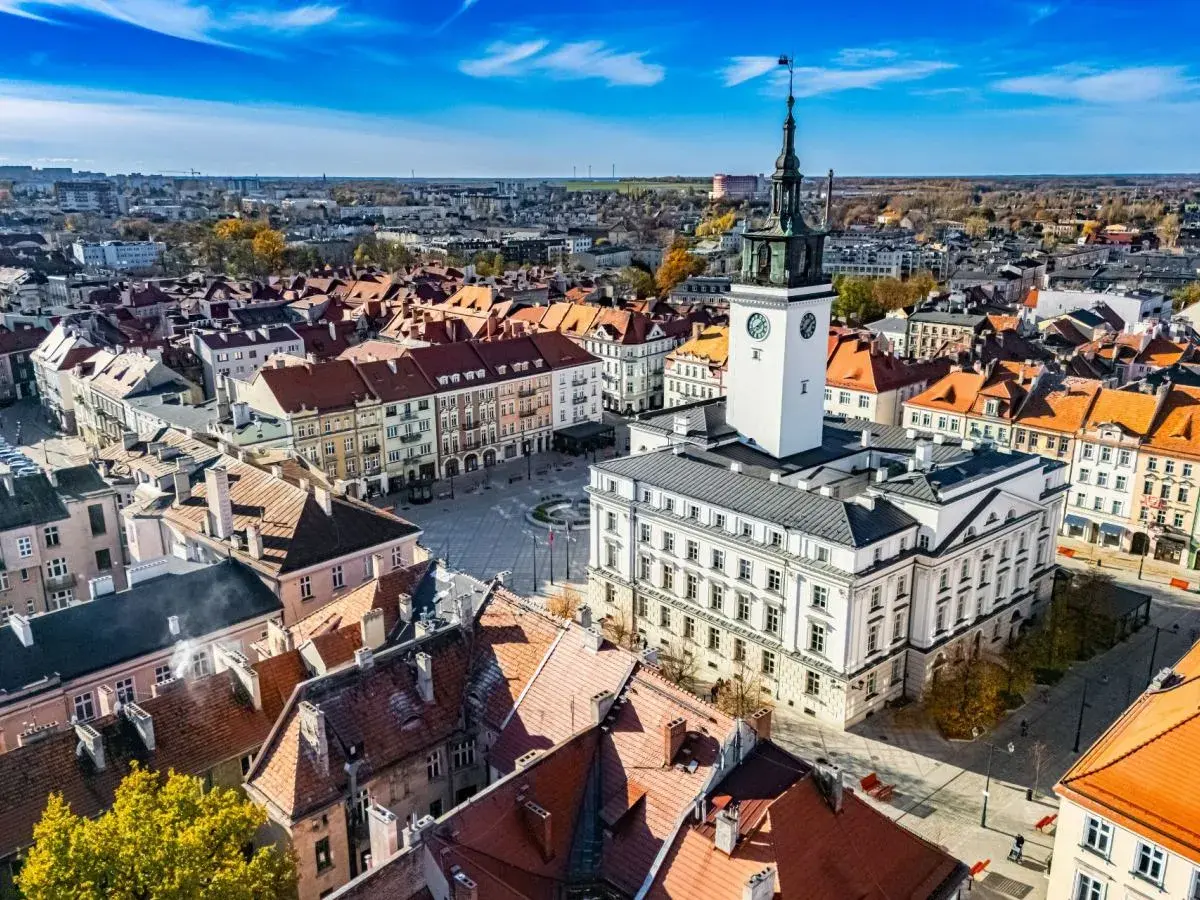 Widok z lotu ptaka na rynek z ratuszem i wieżą zegarową, otoczony kamienicami.