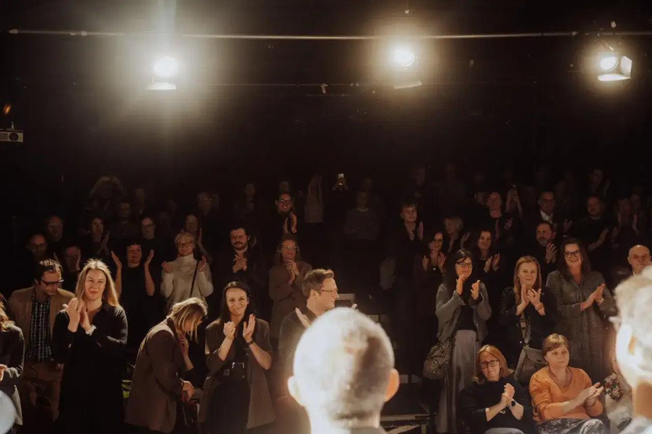 Widownia klaszcze po udanej performance. Ludzie w różnym wieku, oświetleni reflektorami, wyrażają swoje uznanie.